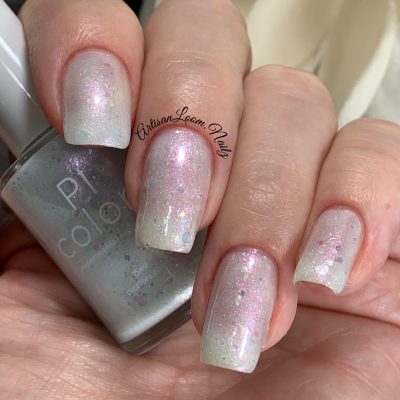 A close-up shot of a hand displays pastel pink and white crelly polish with iridescent flakies for a subtle sparkle, holding a bottle of PI Colors' handmade nail polish, Troth of Innocence.001 White Nail Polish.