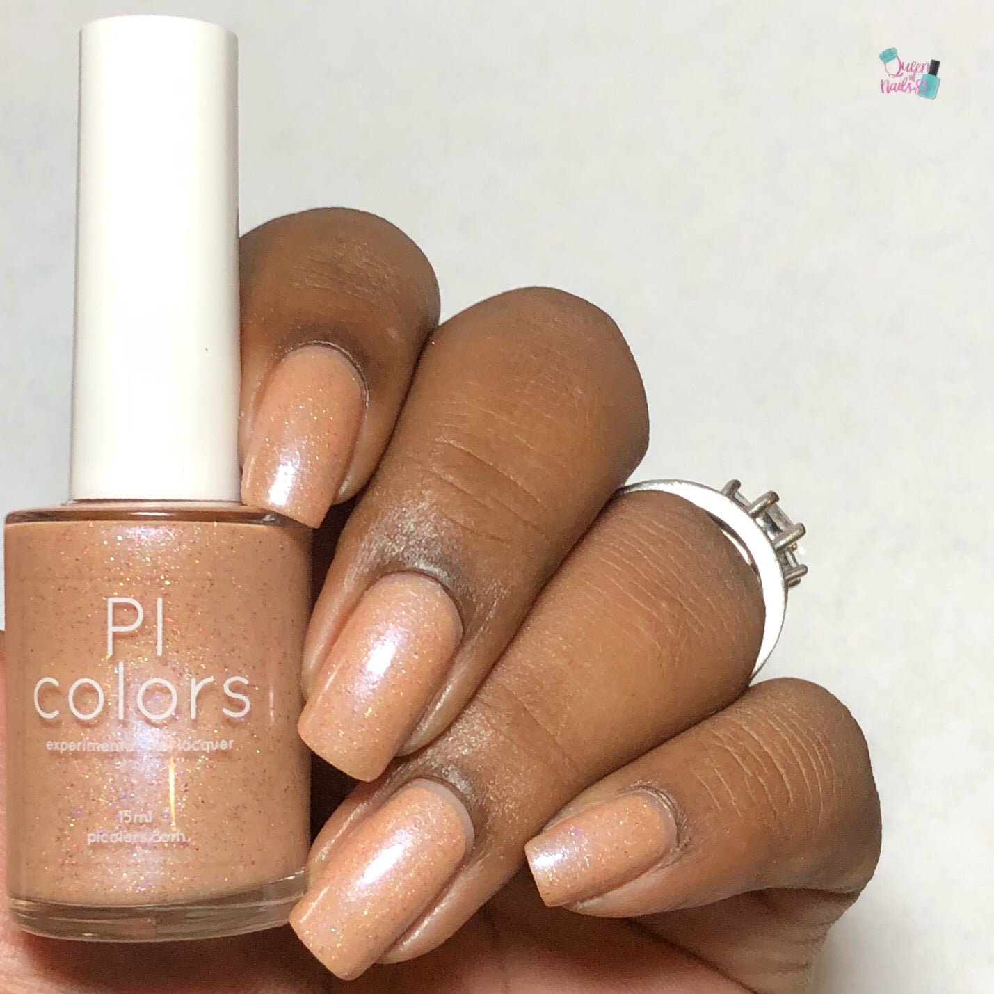 A hand with neatly manicured nails painted in a shimmery nude shade holds a bottle of PI Colors' Sunrise Tellin.027 Pale Orange Nail Polish. The subtle holographic glitter captures the light, adding an extra sparkle against the plain, light backdrop.