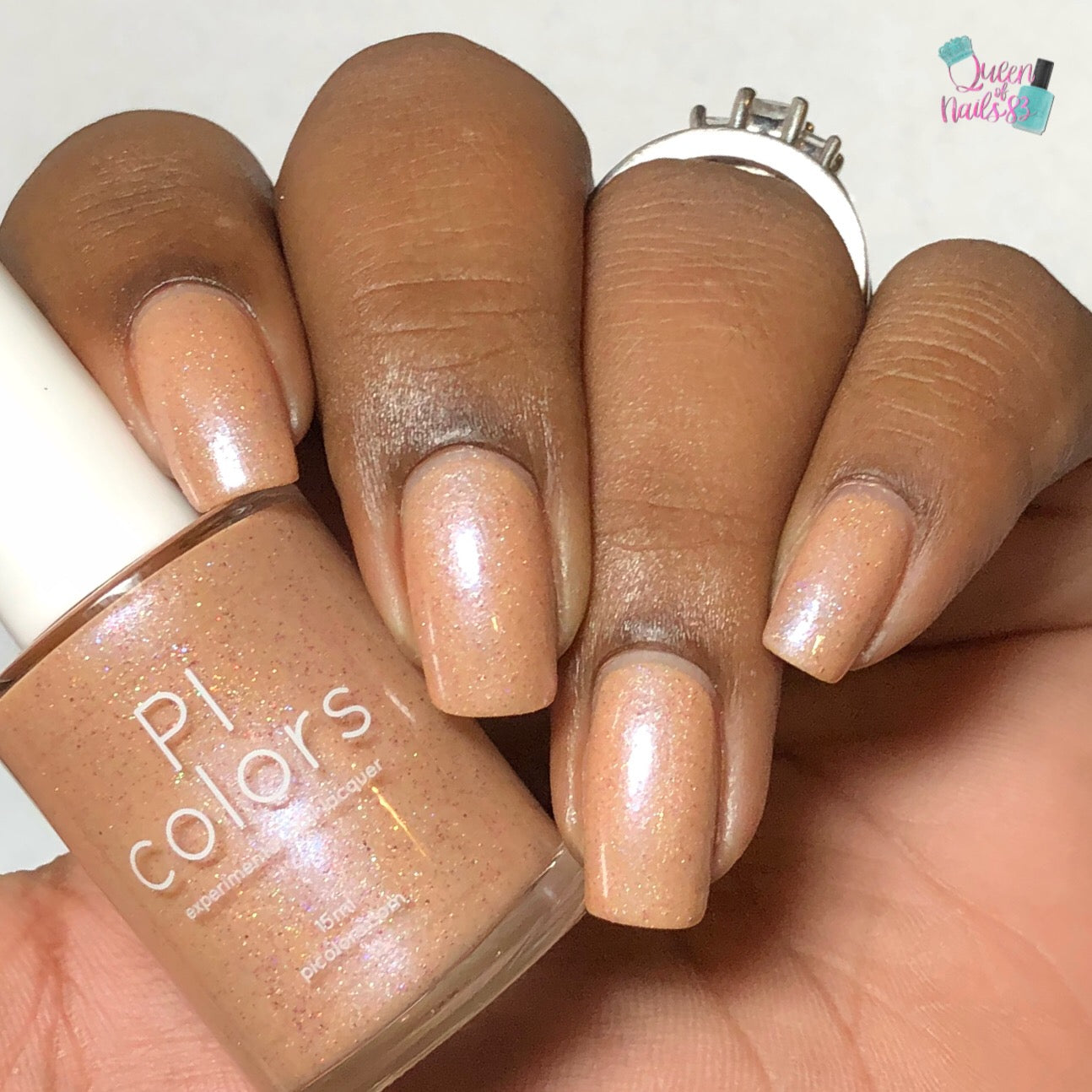 A hand displays neatly manicured nails painted with a shimmery pale orange polish, reminiscent of the shades found in Sunrise Tellin.027 by PI Colors. A bottle with the "PI Colors" label sits in the hand, matching perfectly with the nail color, as holographic glitter introduces a subtle sparkle to the finish. One finger is adorned with a ring featuring an intricate design.