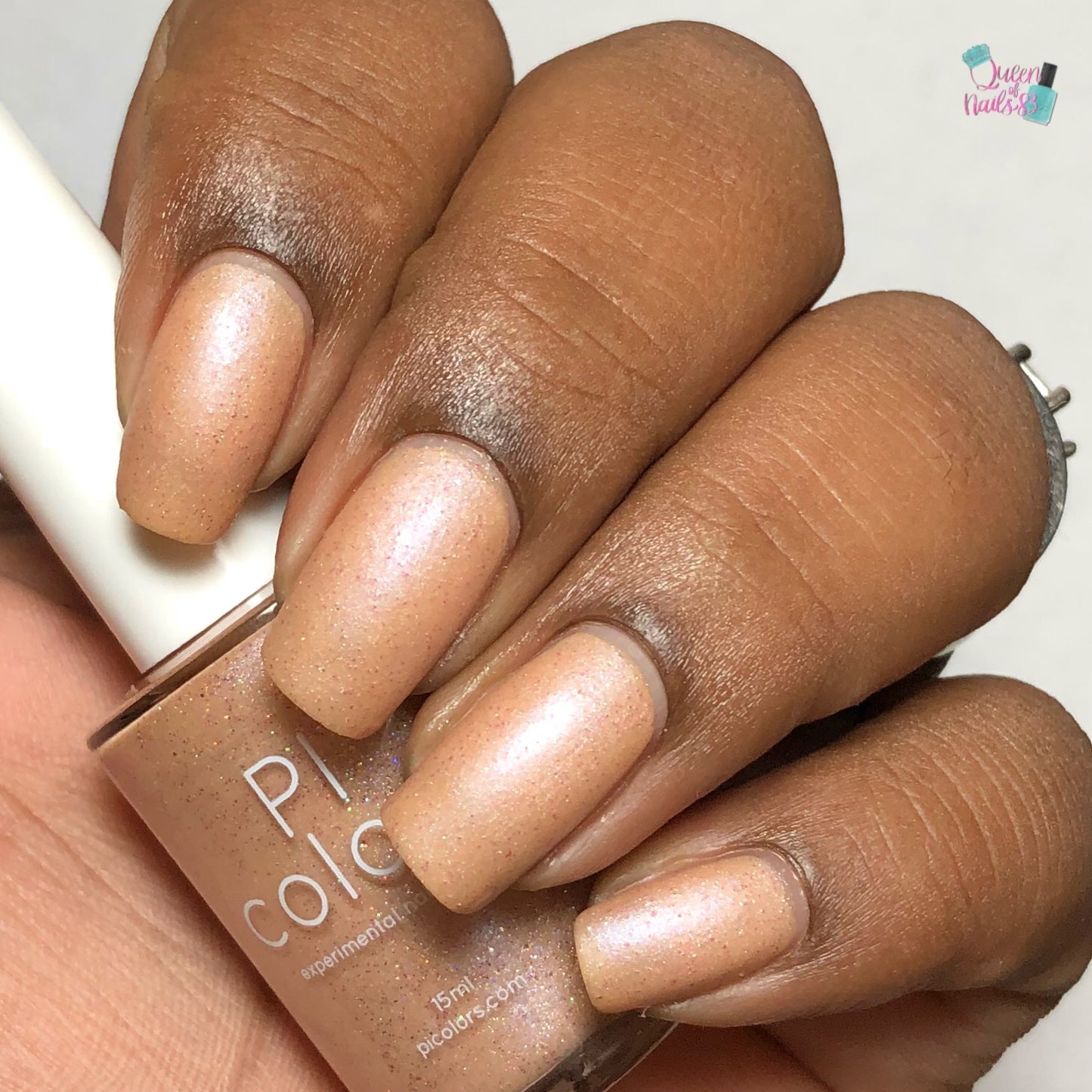 A close-up shows a hand with neatly manicured nails adorned in a shimmery peachy-orange polish that subtly catches the light. The PI Colors Sunrise Tellin.027 Pale Orange Nail Polish bottle is elegantly held between the fingers, partly visible in the frame.