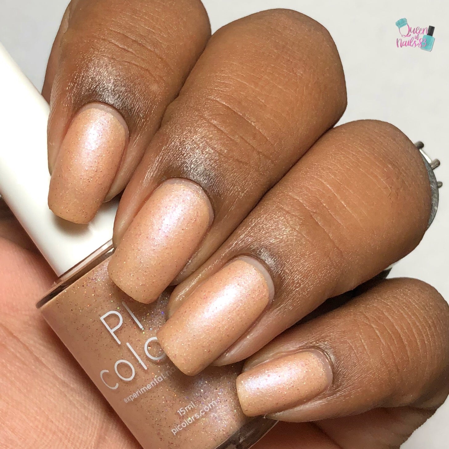 A close-up shows a hand with neatly manicured nails adorned in a shimmery peachy-orange polish that subtly catches the light. The PI Colors Sunrise Tellin.027 Pale Orange Nail Polish bottle is elegantly held between the fingers, partly visible in the frame.
