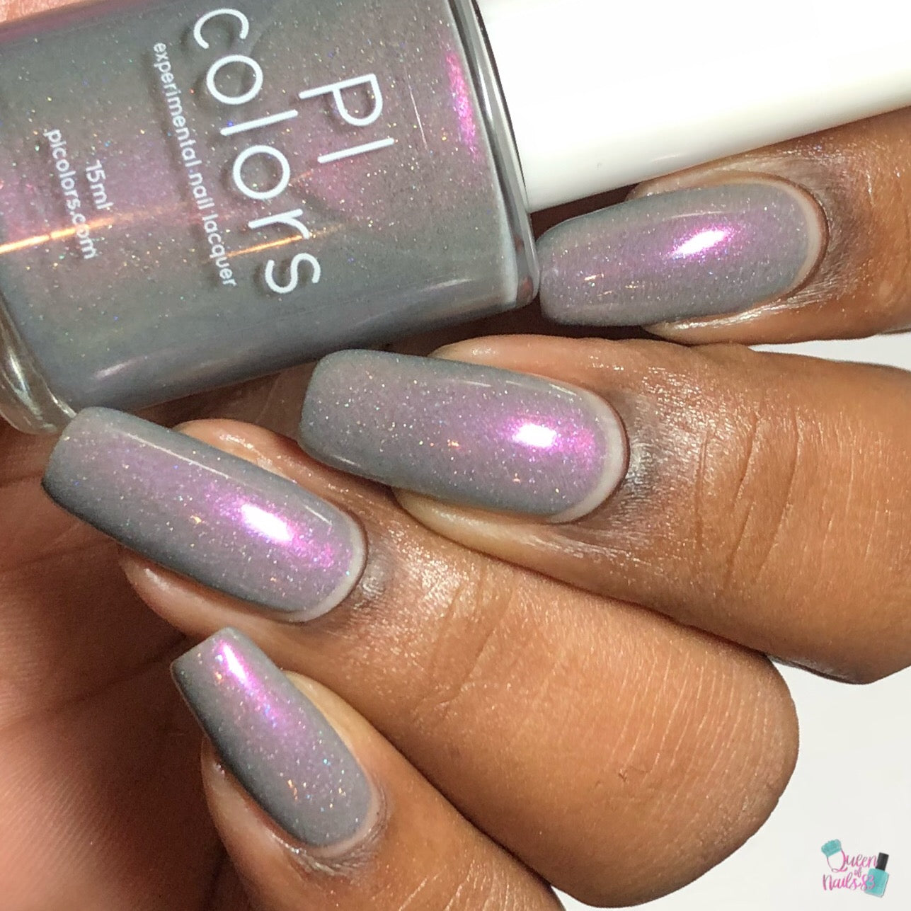 Close-up of a hand with long nails painted in Cirrus.118 Pink Gray nail polish, showcasing a stunning purple shimmer. A bottle of this 10-free nail polish rests between the fingers, labeled "PI Colors.