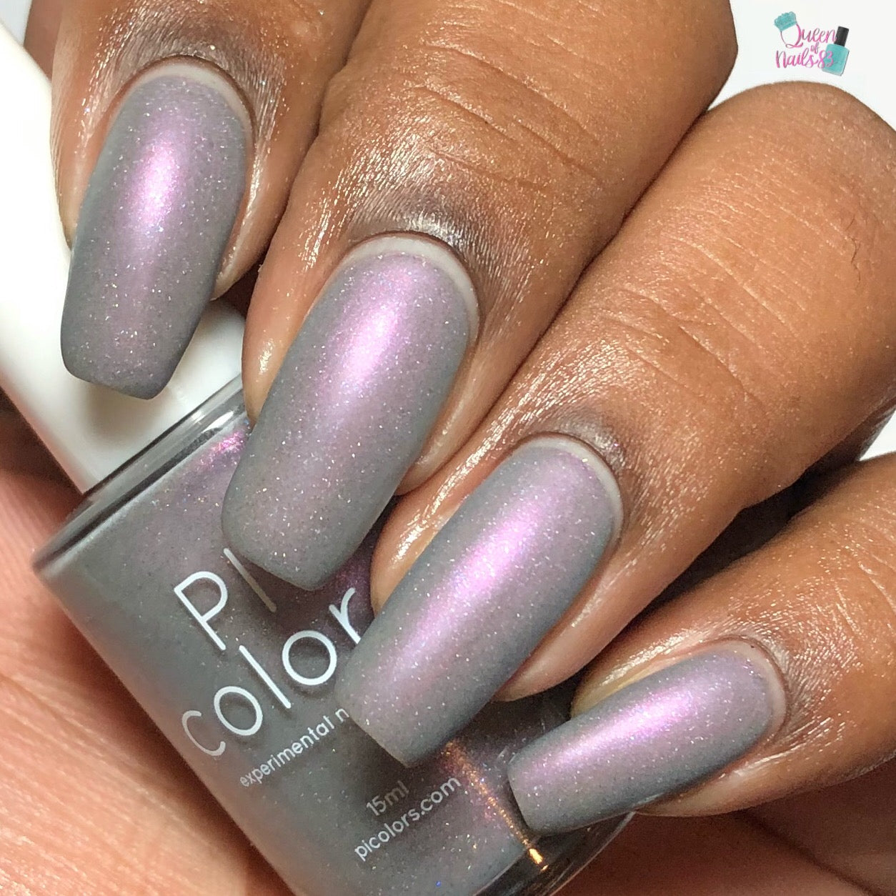 A close-up of a hand showcases gray-based polish from the PI Colors Cirrus.118 collection, featuring a purple shimmer that shifts to pink under light. The long, well-manicured nails complement the matching bottle held nearby, which highlights its 10-free formula with partially visible brand text.