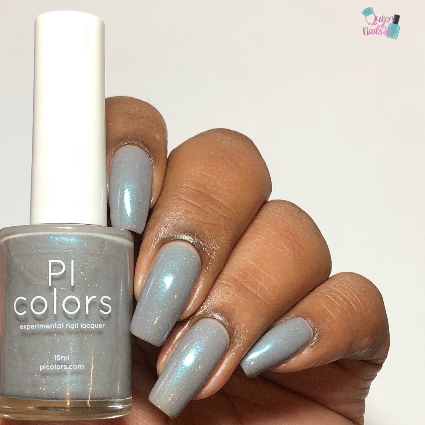 A hand with medium-dark skin tone displays long nails painted in Asperitas.105 blue gray nail polish featuring a hint of holographic sparkle. The thumb and fingers cradle a bottle of PI Colors, set against a plain, light-colored background that highlights the handmade charm.