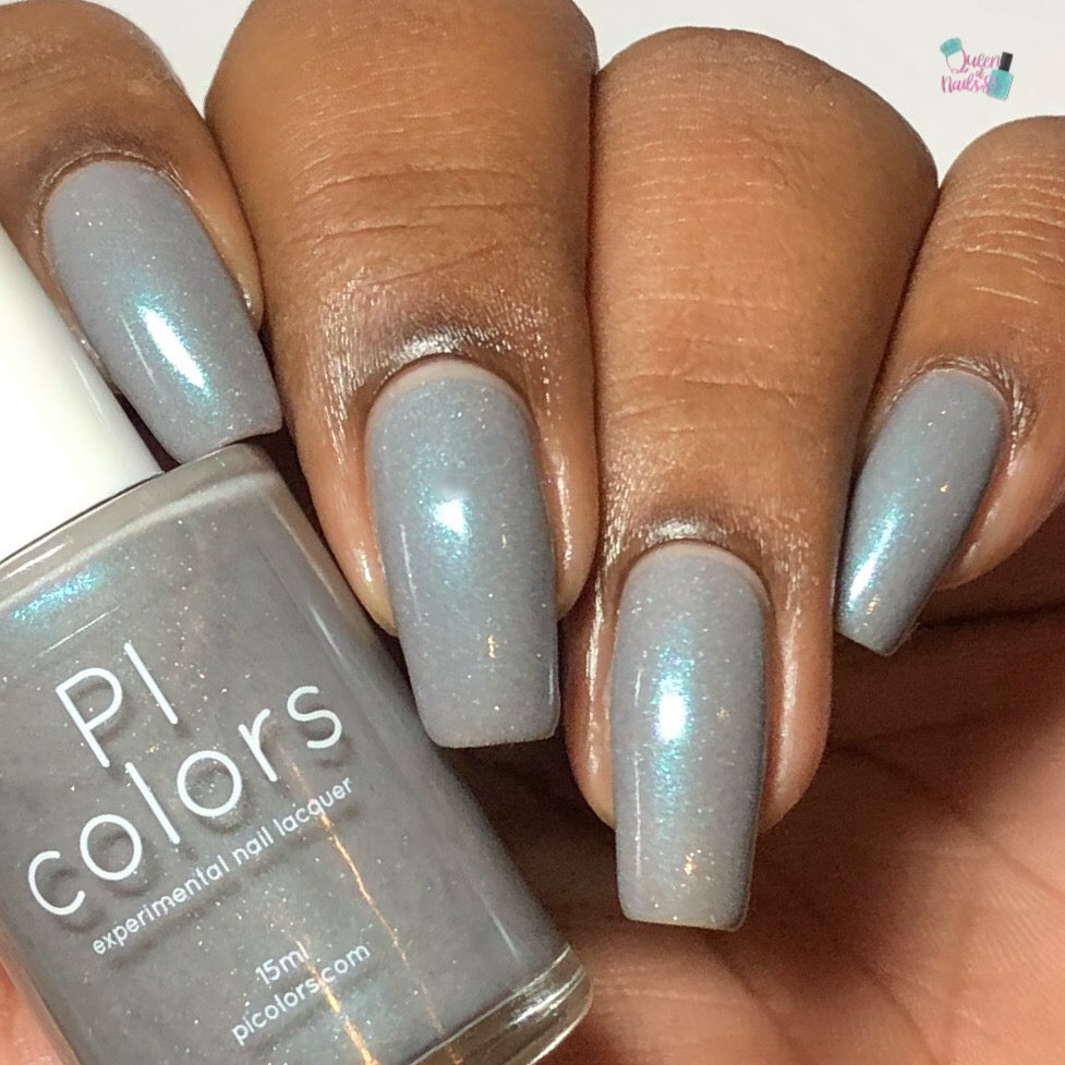 A hand with long fingernails painted in shimmery blue-gray nail polish, infused with holographic sparkle, holds a bottle labeled "Asperitas.105 Blue Gray Nail Polish" by PI Colors. The polish bottle mirrors the dazzling nails perfectly.