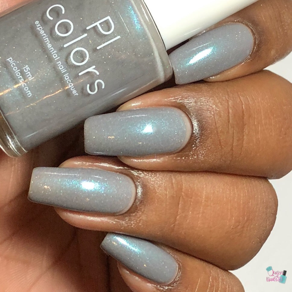 Close-up of a hand with neatly manicured nails painted in a glossy blue gray polish. The bottle of Asperitas.105 Blue Gray Nail Polish is held between the fingers, labeled "PI Colors.