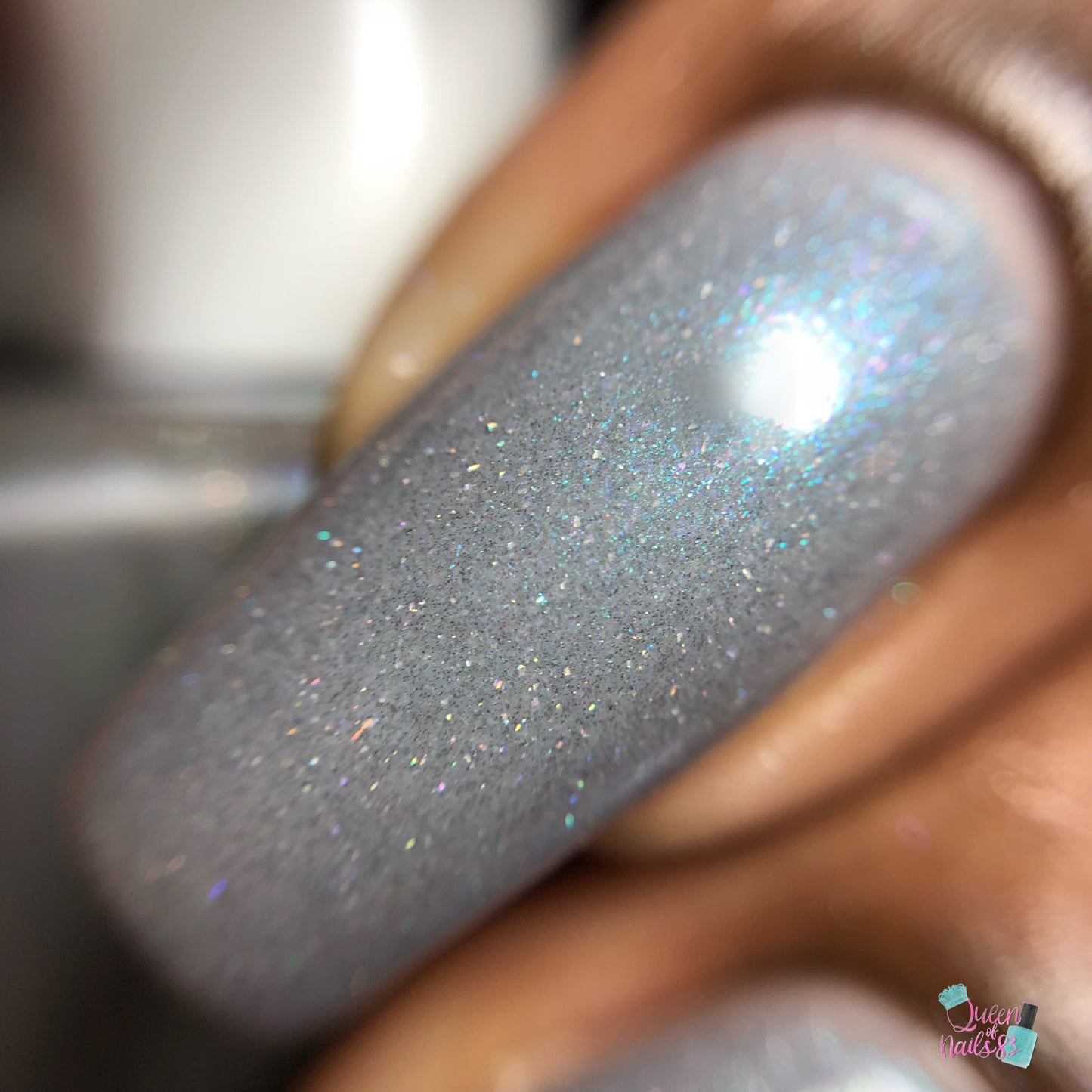 A close-up shot of a nail adorned with Asperitas.105 Blue Gray Nail Polish by PI Colors, shimmering in the light. The surface is smooth and moderately glossy, with small holographic sparkles scattered throughout.