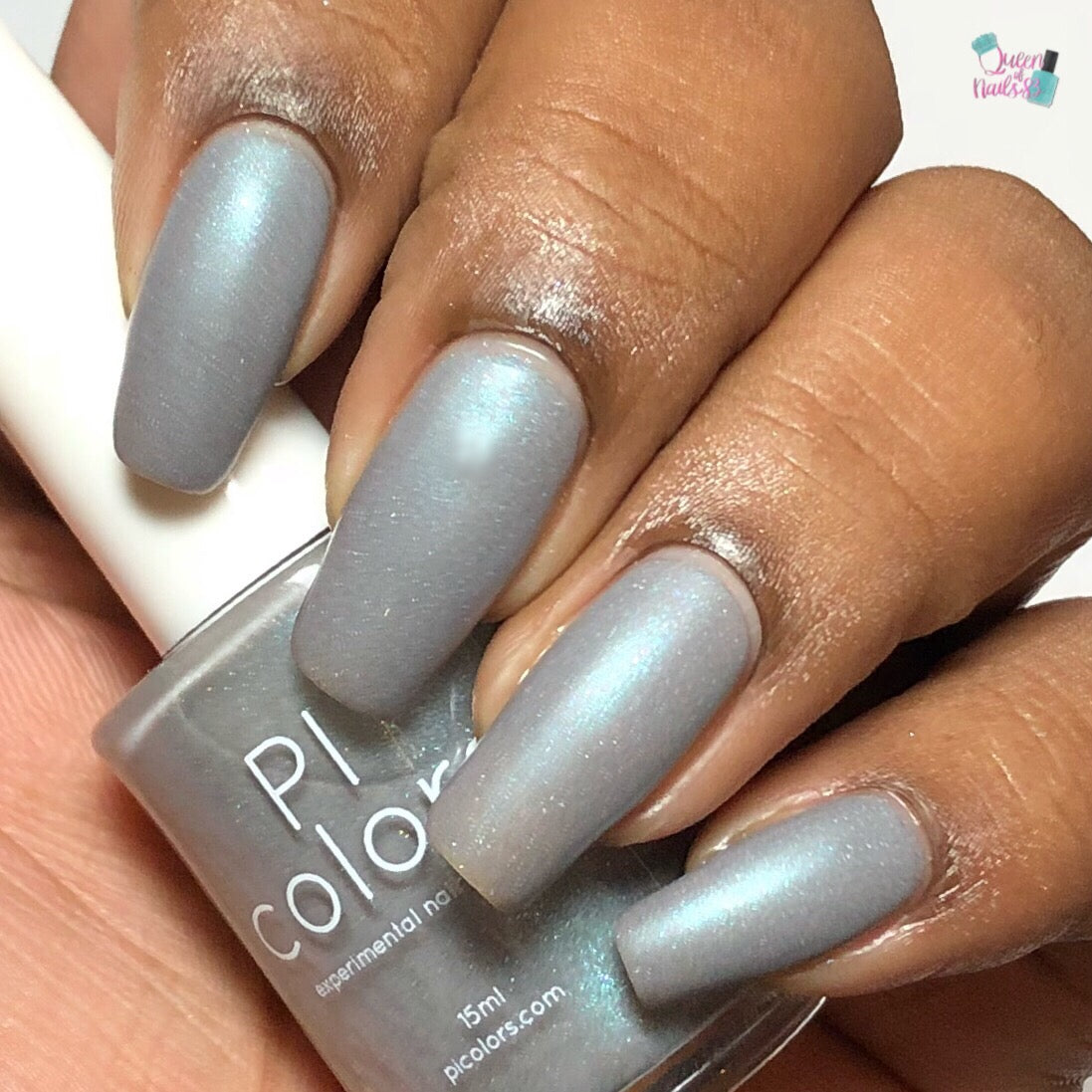 A close-up shot captures a hand with glossy nails painted in PI Colors' Asperitas.105 Blue Gray Nail Polish, showcasing a mesmerizing holographic sparkle as it catches the light. The nails are long and well-manicured, elegantly holding the polish bottle between the fingers.