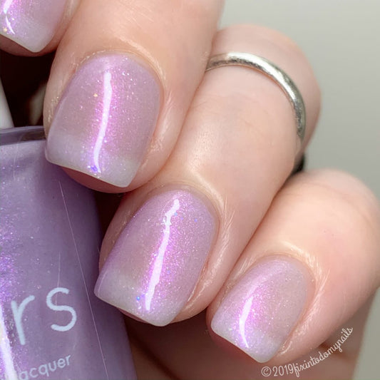 A close-up of a hand with short, neatly manicured nails showcases a 10-free shimmery lavender hue from PI Colors, named Black Cherry Rainbow.200 Purple Nail Polish. The pastel purple polish gleams with a glossy finish, accentuated by rainbow flakies, and the person adorns their middle finger with a simple silver ring.