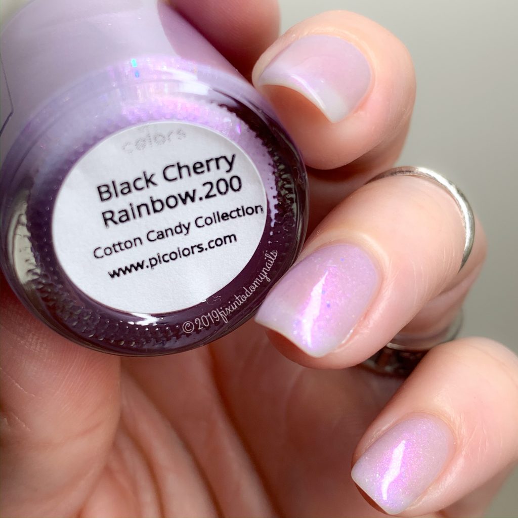 Close-up of a hand holding a PI Colors nail polish bottle labeled "Black Cherry Rainbow.200 Purple Nail Polish" from the Cotton Candy Collection. The nails feature a soft, shimmering lavender hue with rainbow flakies, providing a pastel purple finish that is also 10-free for healthier nails.
