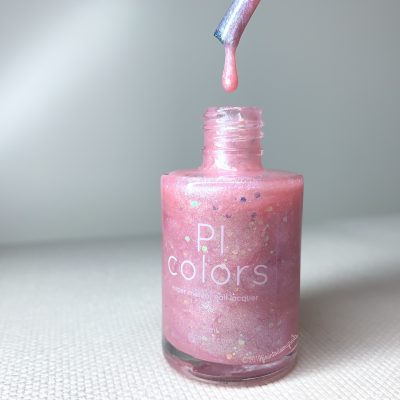 A close-up of a pastel pink jelly nail polish bottle with shimmering particles shows a brush above the bottle, dripping a small drop of polish. The label reads "PI Colors, Pink Candy.210 Luxury Nail Lacquers." The background features a soft, neutral color.
