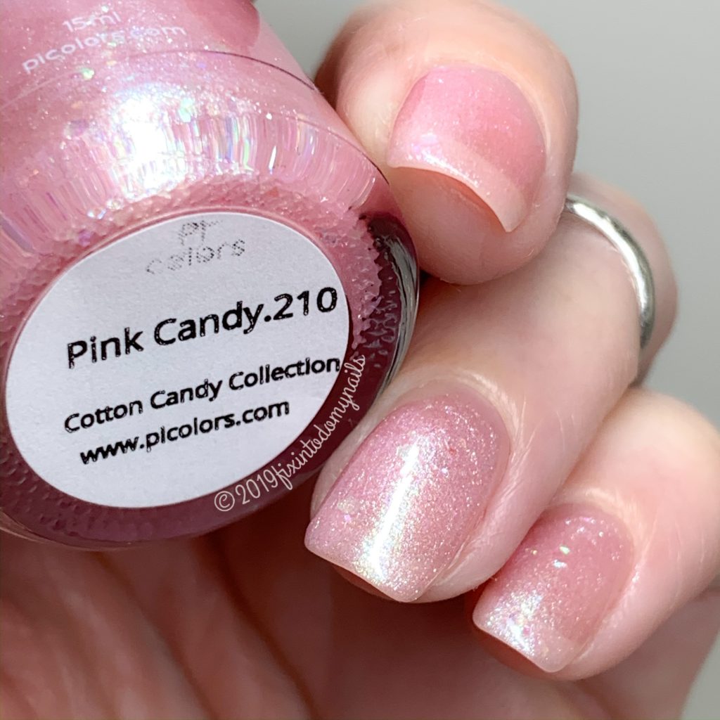 A close-up displays a hand with nails adorned in a shimmery pastel pink jelly polish. The person is holding a bottle of PI Colors' Pink Candy.210 from the Luxury Nail Lacquers Cotton Candy Collection, featuring an elegant and subtle glittery finish.