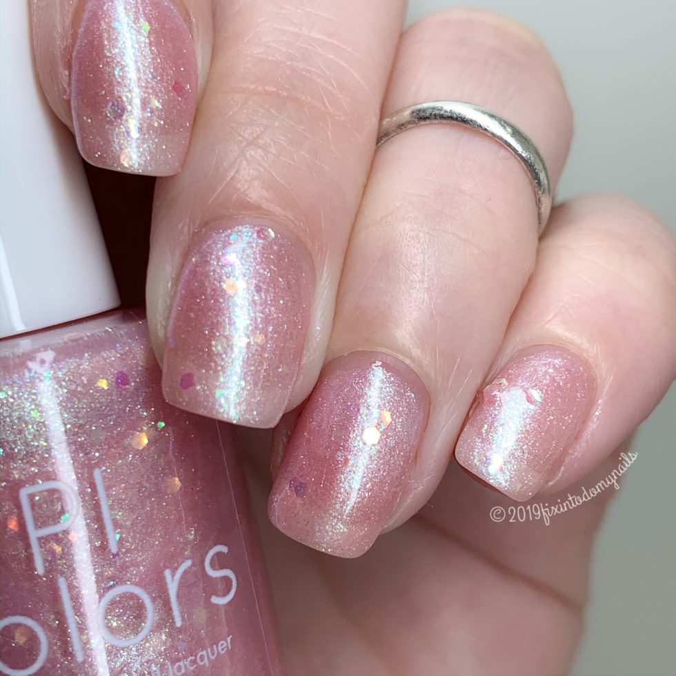 Close-up of a hand with short nails painted in Pink Candy.210 by PI Colors. This luxury nail lacquer features a glittery pastel pink jelly finish, an iridescent shimmer, and subtle heart-shaped glitters. A silver ring adorns one finger.