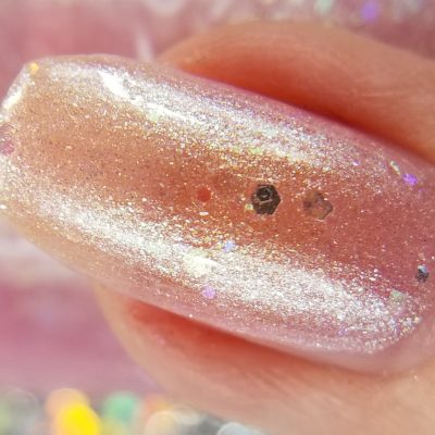 Close-up of a fingernail adorned with PI Colors Pink Candy.210 Nail Polish in pastel pink jelly. The polish features a glossy finish speckled with glitter and small, reflective particles. The softly blurred background reveals hints of pink and multicolored lights reminiscent of the polish's vibrant theme.
