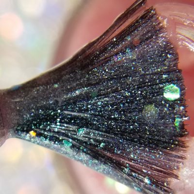 Close-up of a makeup brush dipped in shimmering, multicolored glitter with fine particles of blue, green, and silver. The softly blurred background enhances the sparkling bokeh effect, reminiscent of the pastel pink jelly elegance found in PI Colors' Pink Candy.210 Pink Nail Polish.