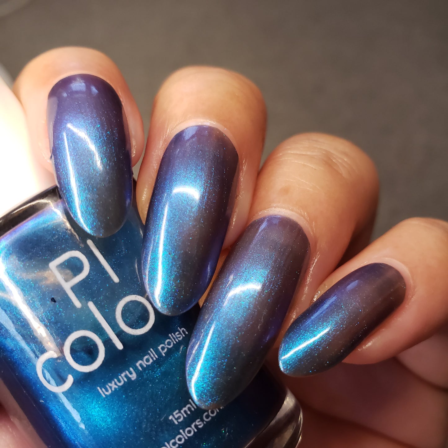 A close-up captures a hand with elegantly long fingernails painted in Aurora Blue.316, showcasing the luxurious and iridescent hue. The handmade nail polish bottle is held in the hand, prominently displaying the "PI Colors" label. The nails exhibit a glossy, metallic sheen that radiates sophistication.