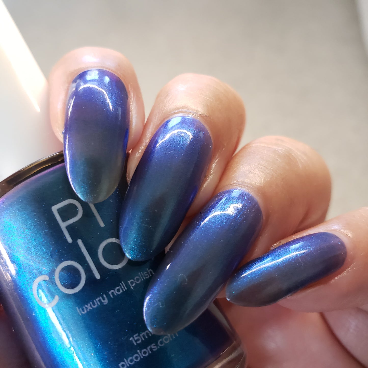 A close-up shot captures a hand with long nails elegantly coated in Aurora Blue.316, a glossy metallic blue-purple luxury nail lacquer. The impeccably manicured nails gleam brilliantly. A portion of the PI Colors bottle is visible, featuring the word "luxury," suggesting its artisanal craftsmanship.