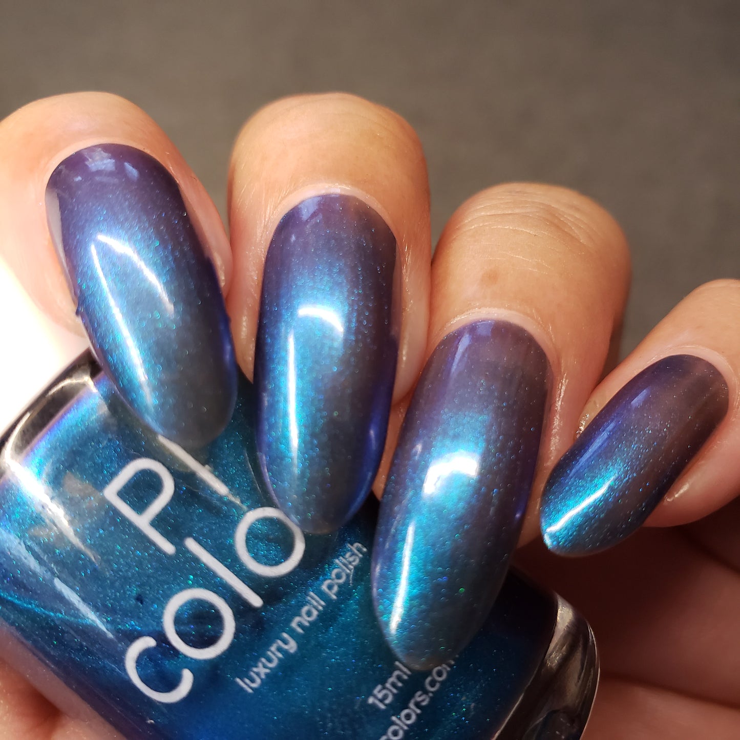 Close-up of a hand with long nails, adorned with PI Colors' luxury nail lacquer in Aurora Blue.316. The shimmering polish transitions to a stunning purple hue under different lighting, while the thumb elegantly holds a bottle of this handcrafted nail polish.