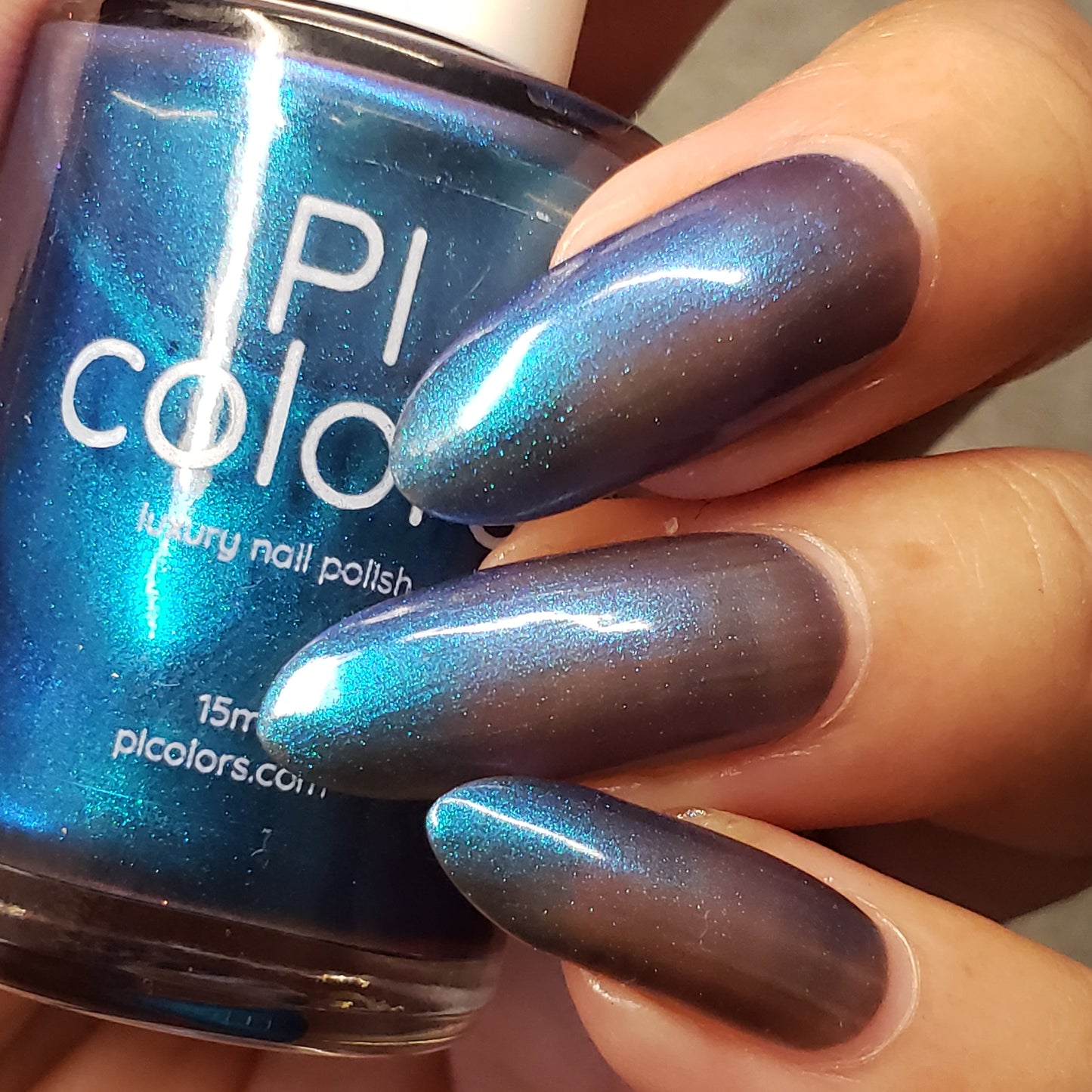 A close-up of a hand with long nails painted in the luxurious, iridescent Aurora Blue.316 is seen holding a bottle of handmade nail polish labeled with the brand name "PI Colors." This exquisite lacquer transitions between striking blue and purple shades when exposed to light.