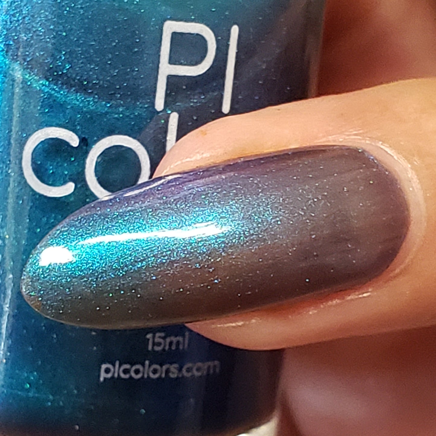 A close-up captures a hand holding a bottle of PI Colors' luxury nail lacquer. The polished nail displays a striking metallic gradient from Aurora Blue.316 to purple, with a glossy finish. Partially visible behind it, the bottle features a blue color adorned with white text.