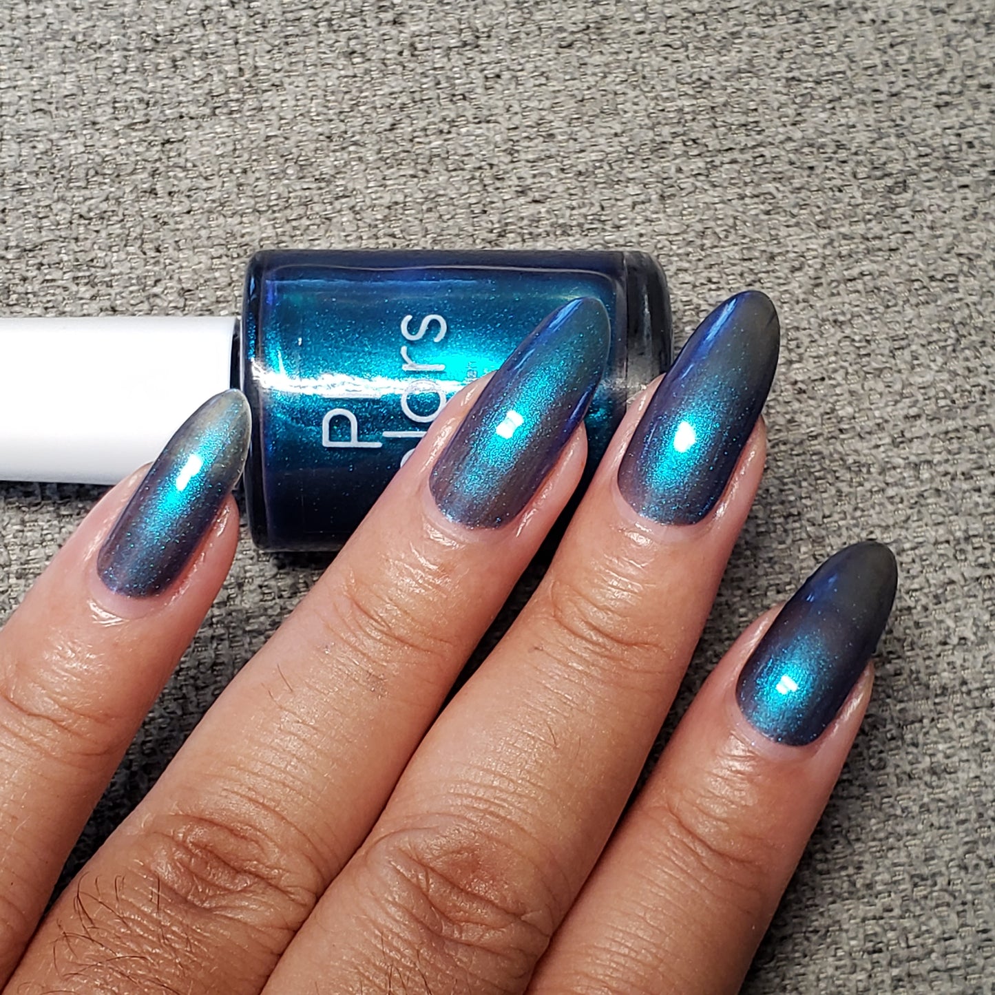 A hand with long, pointed nails painted in a luxurious PI Colors nail lacquer named Aurora Blue.316 rests on a gray textured surface. A matching bottle of this handmade nail polish is placed above the hand, completing the elegant display.