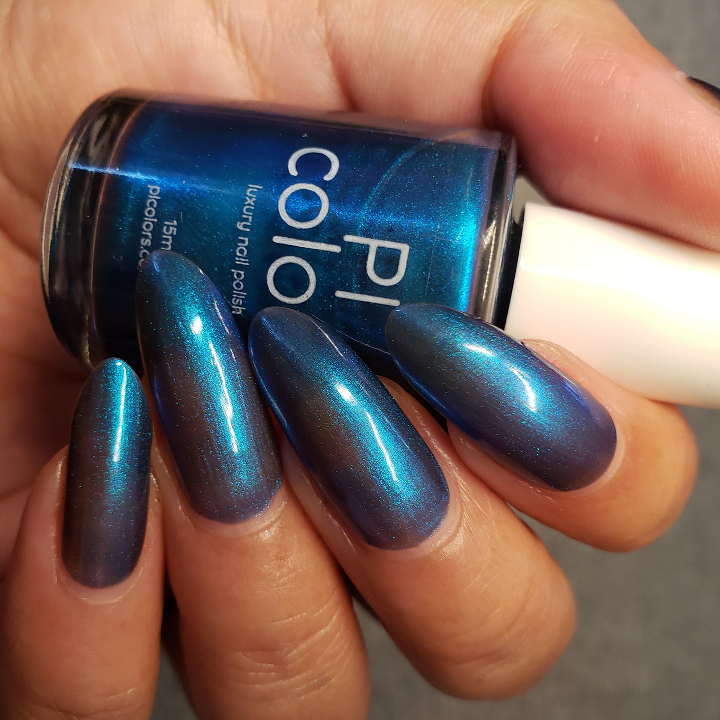 A close-up shot showcases a hand with long, oval-shaped nails adorned in a shimmering electric blue polish. The hand is holding a matching bottle of PI Colors' Aurora Blue.316 nail polish, featuring a white cap and promoting its 10-free formula for safer beauty choices.