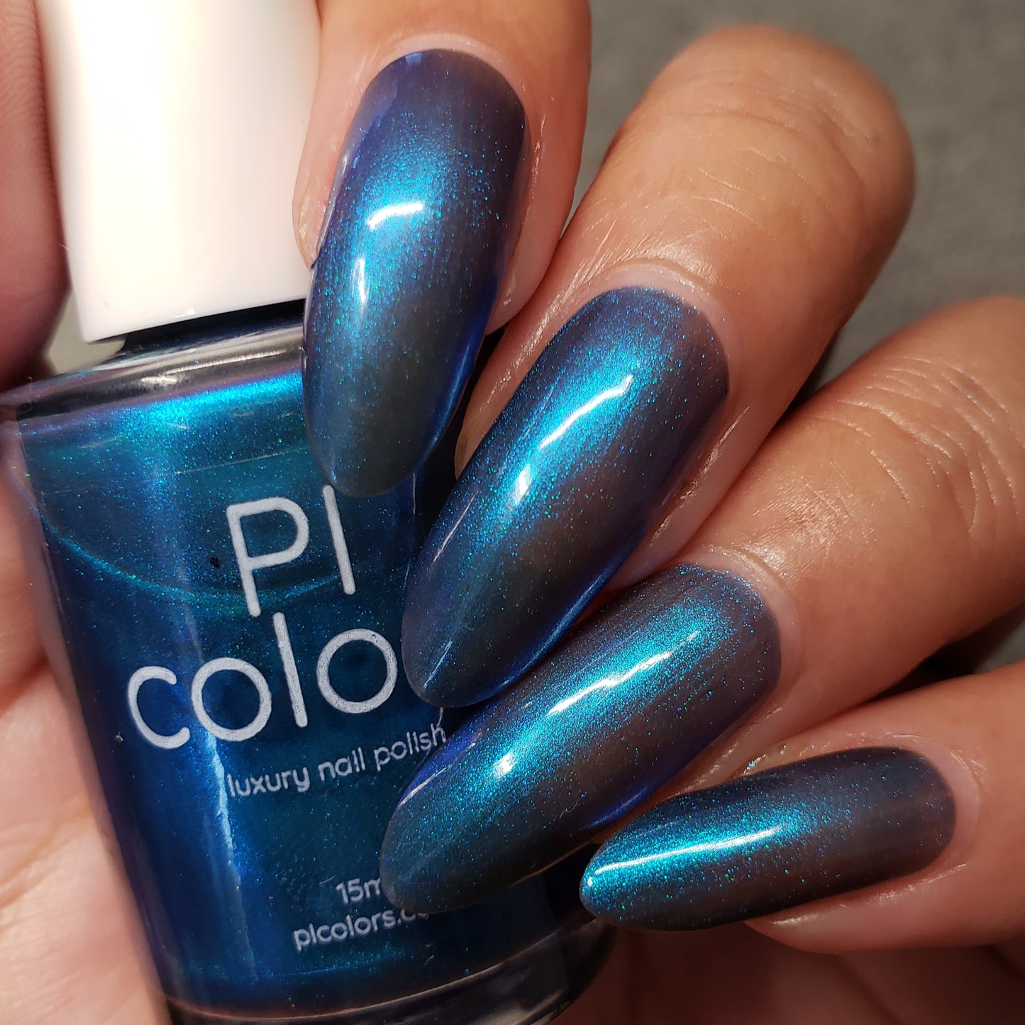 Close-up of a hand with long nails painted in Aurora Blue.316 nail polish, next to a bottle labeled "PI Colors." The 10-free polish reflects light, creating a shimmery effect.