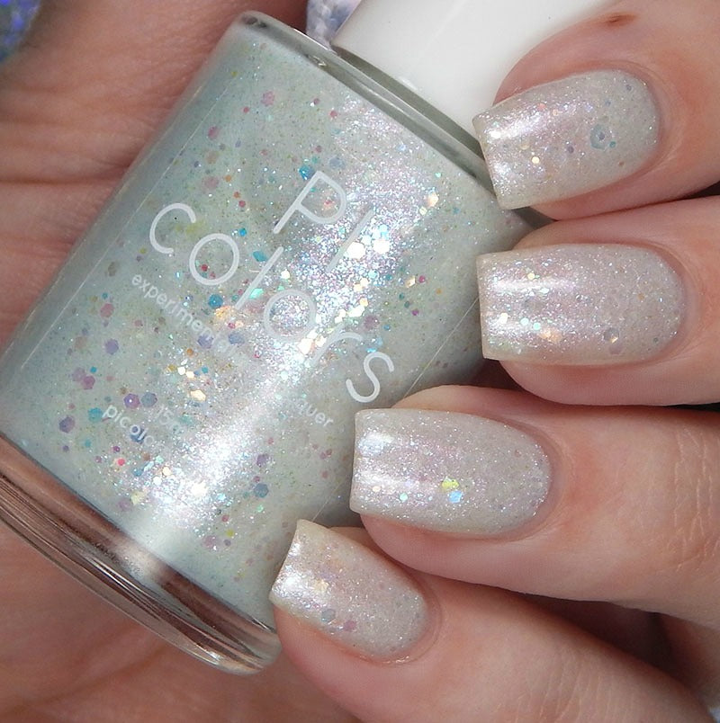 A well-manicured hand, with nails adorned in shimmery pastel hues, holds a bottle of PI Colors' Troth of Innocence.001 White Nail Polish. This handmade polish features a light, glittery finish with multicolored specks and iridescent flakies for extra sparkle.