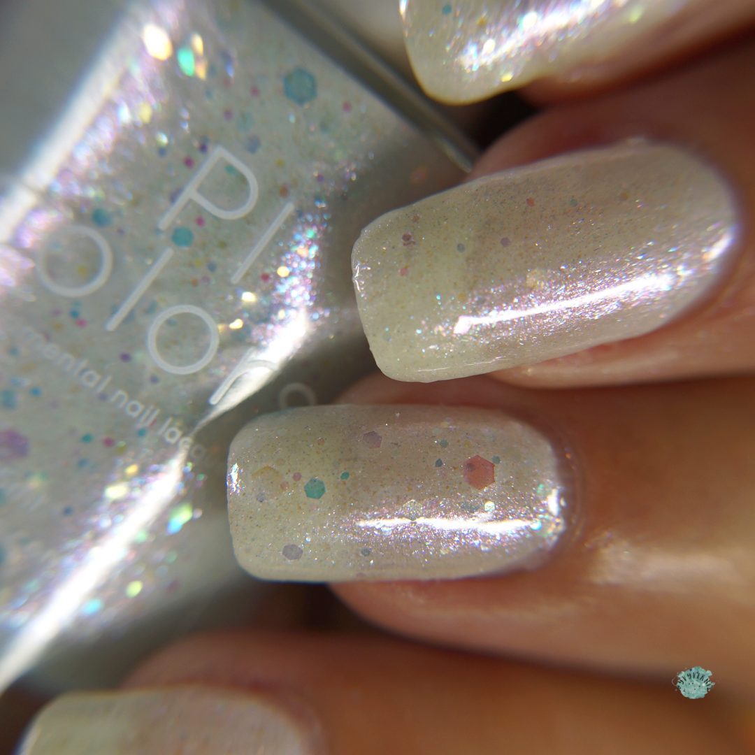 Close-up of fingers adorned with the semi-transparent, shimmering "Troth of Innocence.001" white crelly polish by PI Colors, showcasing iridescent flakies and multicolored glitter. In the background, a bottle of this matching handmade nail polish is partially visible.