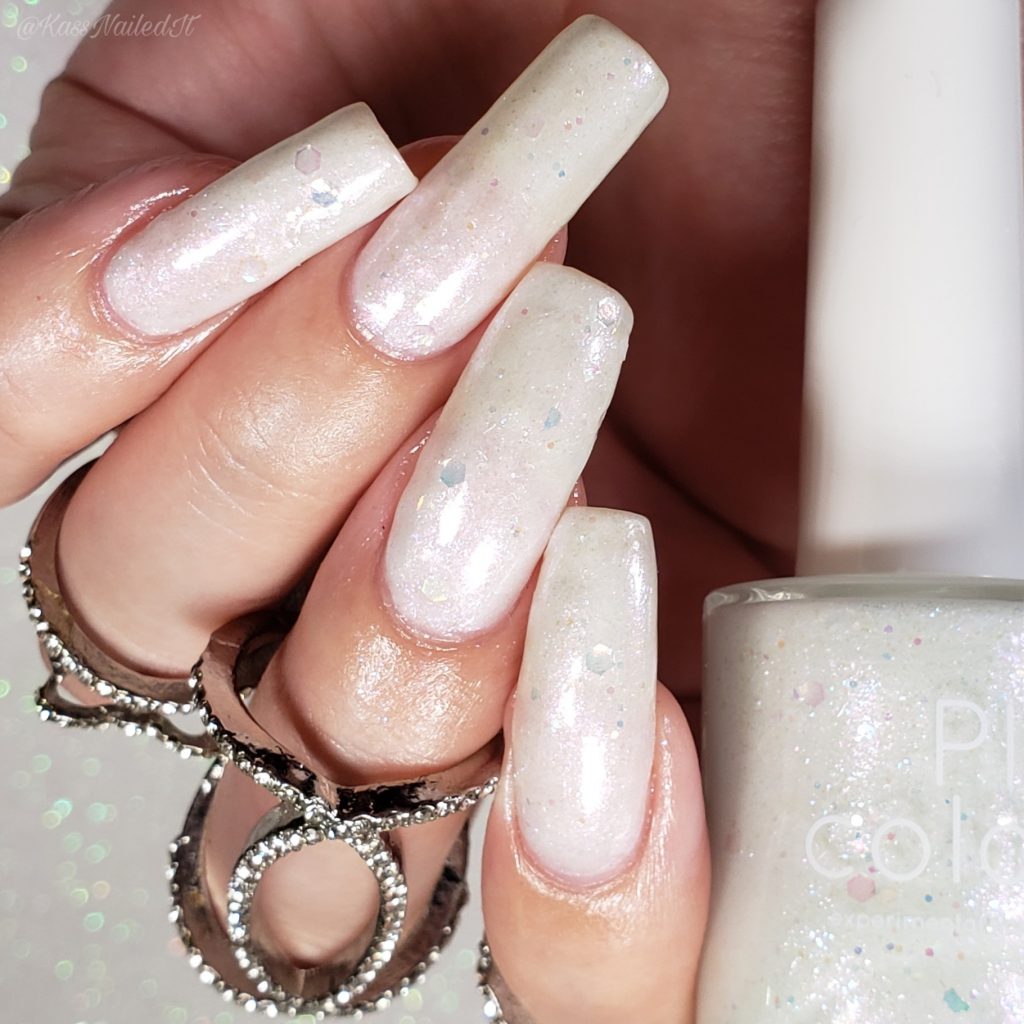 A close-up captures a hand adorned with long nails painted in Troth of Innocence.001 White Nail Polish by PI Colors, shimmering with iridescent flakies. The person wears a silver ring on their finger against a sparkly background, emphasizing the polish's glittery elegance.