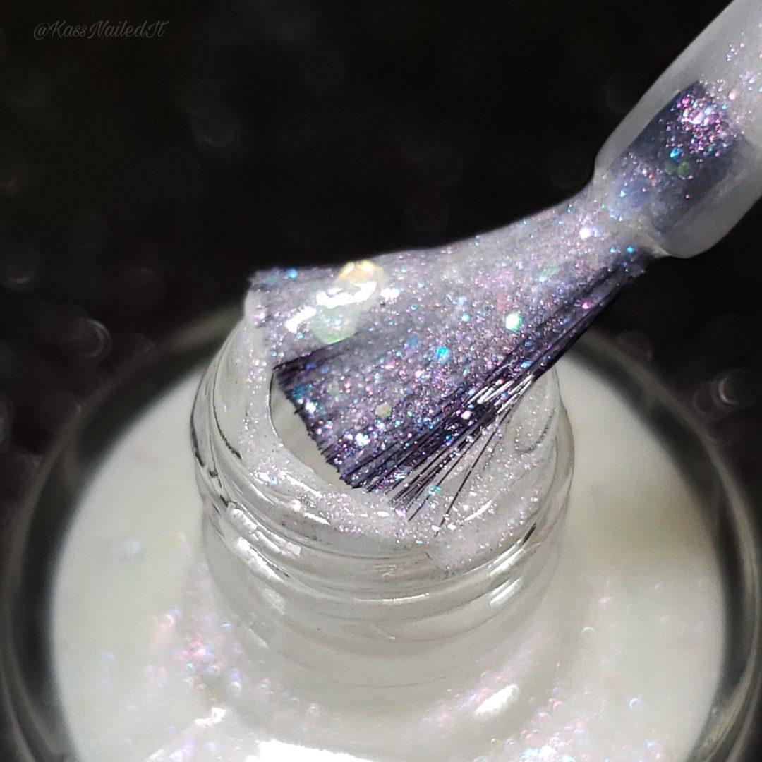 Close-up of a nail polish brush coated in Troth of Innocence.001 by PI Colors, a glittery, purple-tinted handmade nail polish being pulled from the bottle. The polish sparkles with tiny multicolored specks and iridescent flakies.