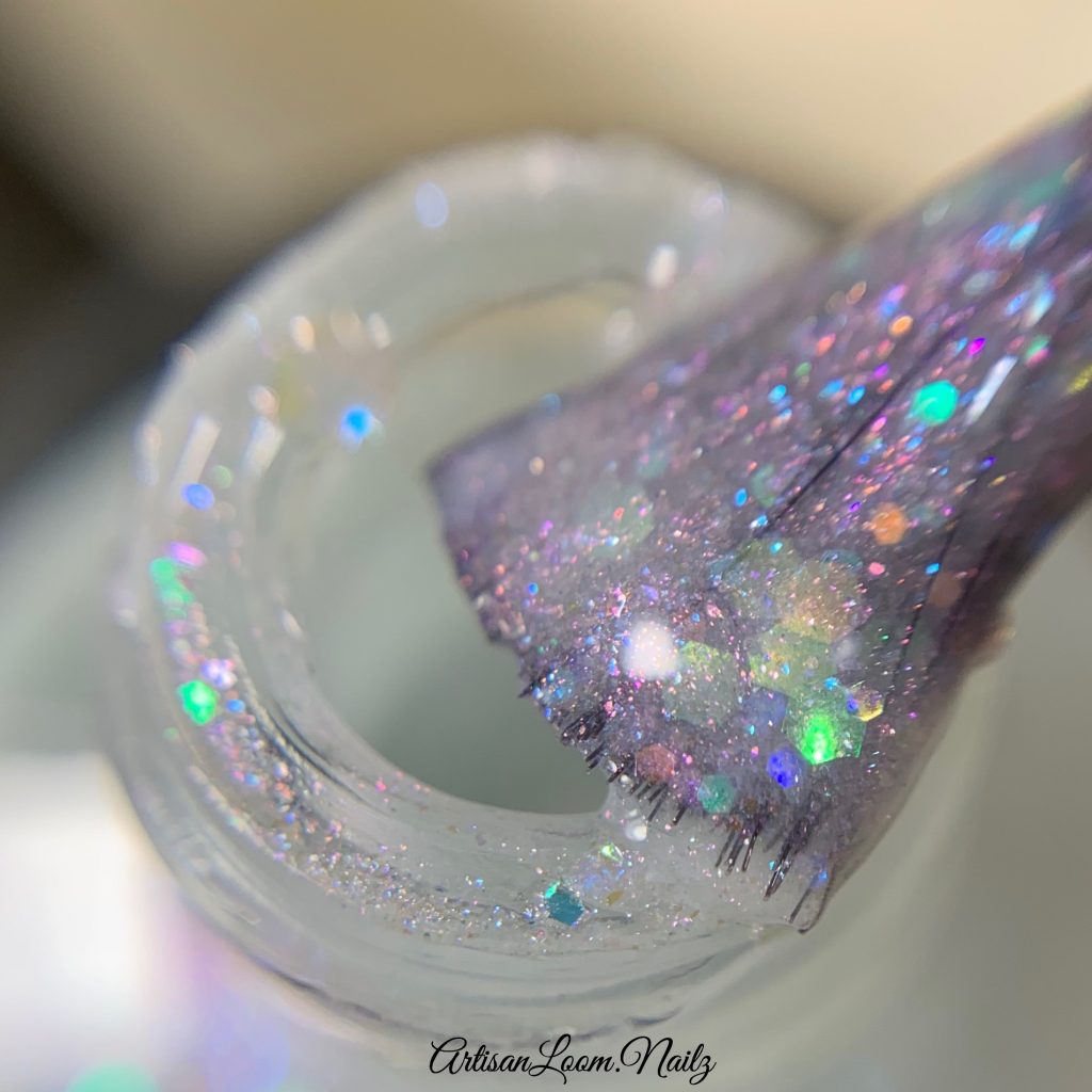 A close-up of a brush dipped in a jar of PI Colors' Troth of Innocence.001 White Nail Polish, which is handmade and adorned with glittery, iridescent flakies. This polish sparkles with an array of colors under the light, creating a shimmering effect.