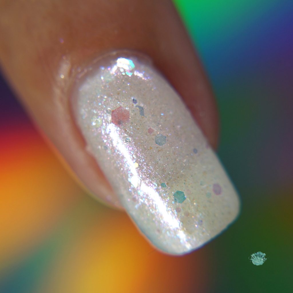 Close-up of a fingernail adorned with PI Colors' "Troth of Innocence.001" white nail polish, a shimmery crelly featuring small colorful specks. The background showcases a blurred array of rainbow hues, highlighting the charm of the iridescent flakies within this handmade polish.