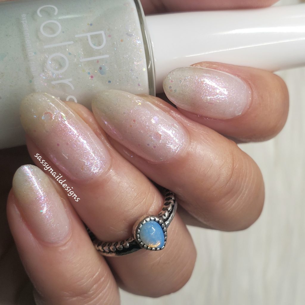 A hand with neatly manicured nails painted in the shimmery, gradient pastel hue of PI Colors' Troth of Innocence.001 White Nail Polish is shown. The ring finger flaunts a silver ring with a blue stone. A partially visible bottle of this handmade nail polish adds charm to the scene, while iridescent flakies enhance the ethereal glow.