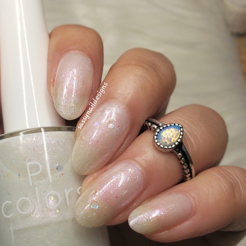 A hand with long, almond-shaped nails painted in sheer, pearlescent finish holds a bottle of Troth of Innocence.001 White Nail Polish by PI Colors. This polish features iridescent flakies, adding a subtle shimmer. The person also wears a ring with an oval opal stone, enhancing the overall elegance.