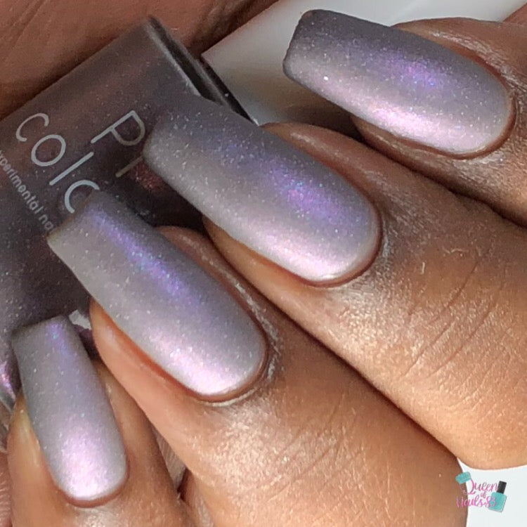 A close-up captures a person's hand adorned with long, matte lavender nails accented by delicate holographic glitter. In the background, the nail polish bottle is partially visible, showcasing its luxury brand name, PI Colors, and proudly displaying "Uranus in Bloom.092 Purple Nail Polish.