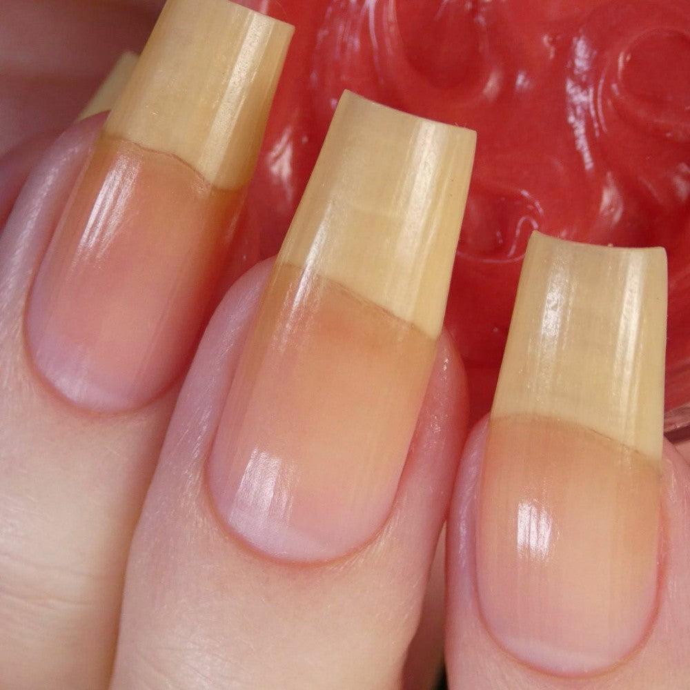 Close-up of long, natural fingernails with a light yellow tint, set against a blurred background of a red textured surface. The nails have a smooth, glossy appearance, enhanced by PI Colors’ Strawberry Scented Cuticle Butter, highlighting their well-maintained condition.