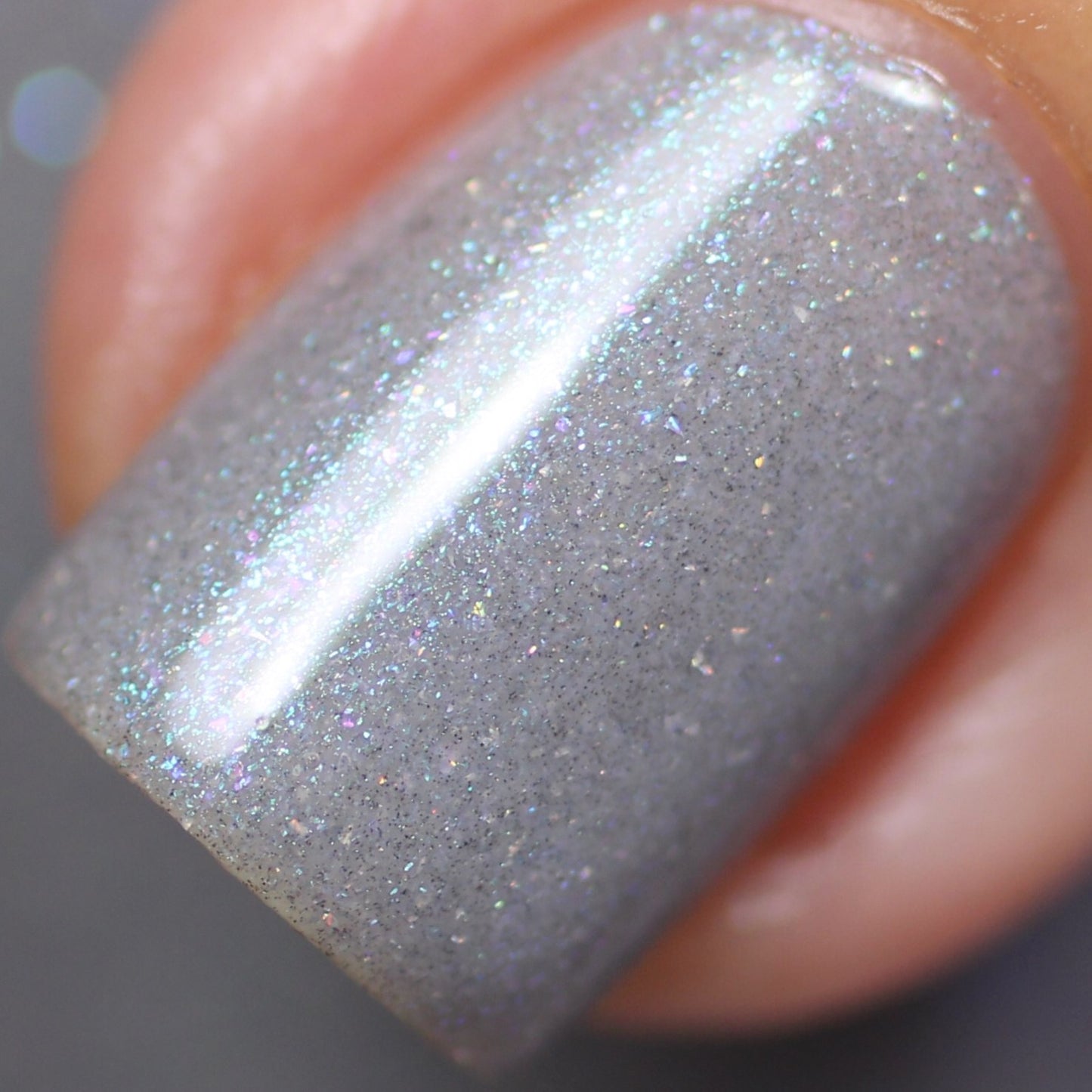 A close-up of a fingernail adorned with PI Colors' Asperitas.105 Blue Gray Nail Polish reveals its shimmery hue. This handmade nail polish flaunts a holographic sparkle, catching various colors when it hits the light. The surrounding skin is subtly visible, signifying a natural nail application.