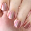 A close-up of a person's hand showcases neatly manicured nails, adorned with the luxurious Beta Ceti.055 Rust Orange Red Nail Polish by PI Colors. The polish's multichrome shimmer captivates the light, emphasizing its gleam and elegance. A softly blurred background enhances the focus on this exquisite nail art.