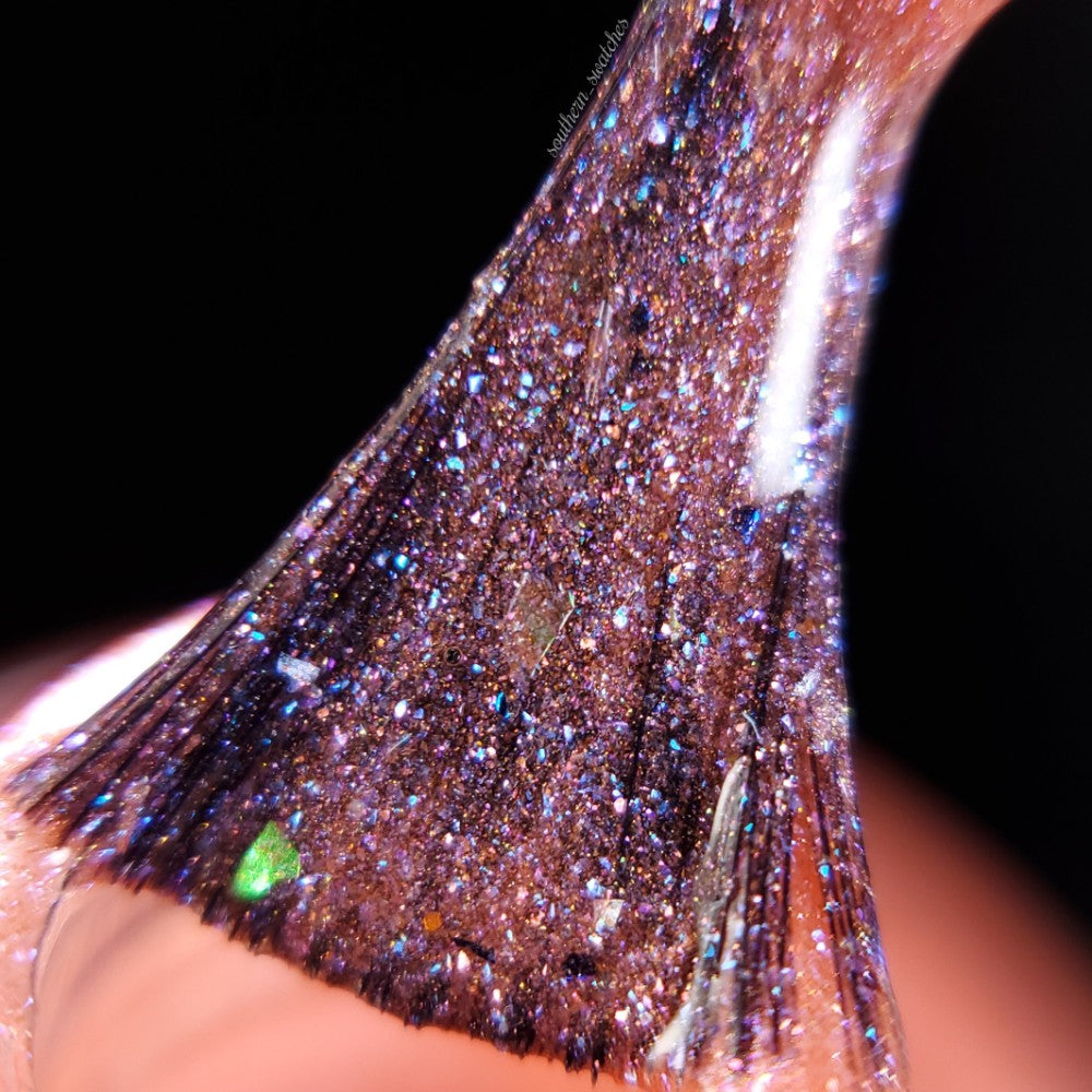 A close-up of the Lala Ngoxolo Kumkani Coral Nail Polish by PI Colors shows a glossy, glittery substance being stretched. The material sparkles with shades of purple and blue, similar to iridescent rainbow flakes, reflecting light and highlighting its textured surface against a dark background.