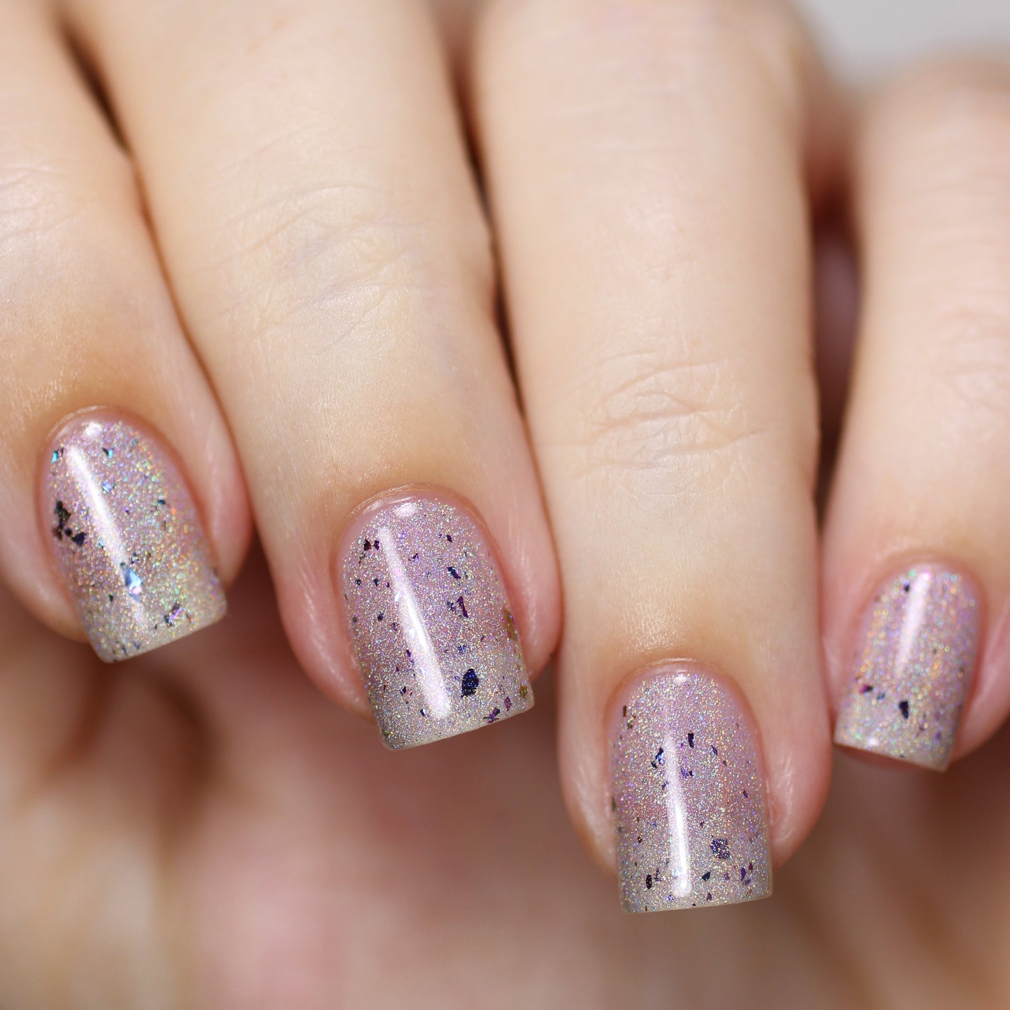 A close-up of a hand with nails painted in PI Colors' Crescent in Ice.018 Silver Nail Polish, featuring a shimmering silvery lilac hue accented by black speckles. The well-manicured nails have a glossy finish, highlighting a trendy and artistic style.