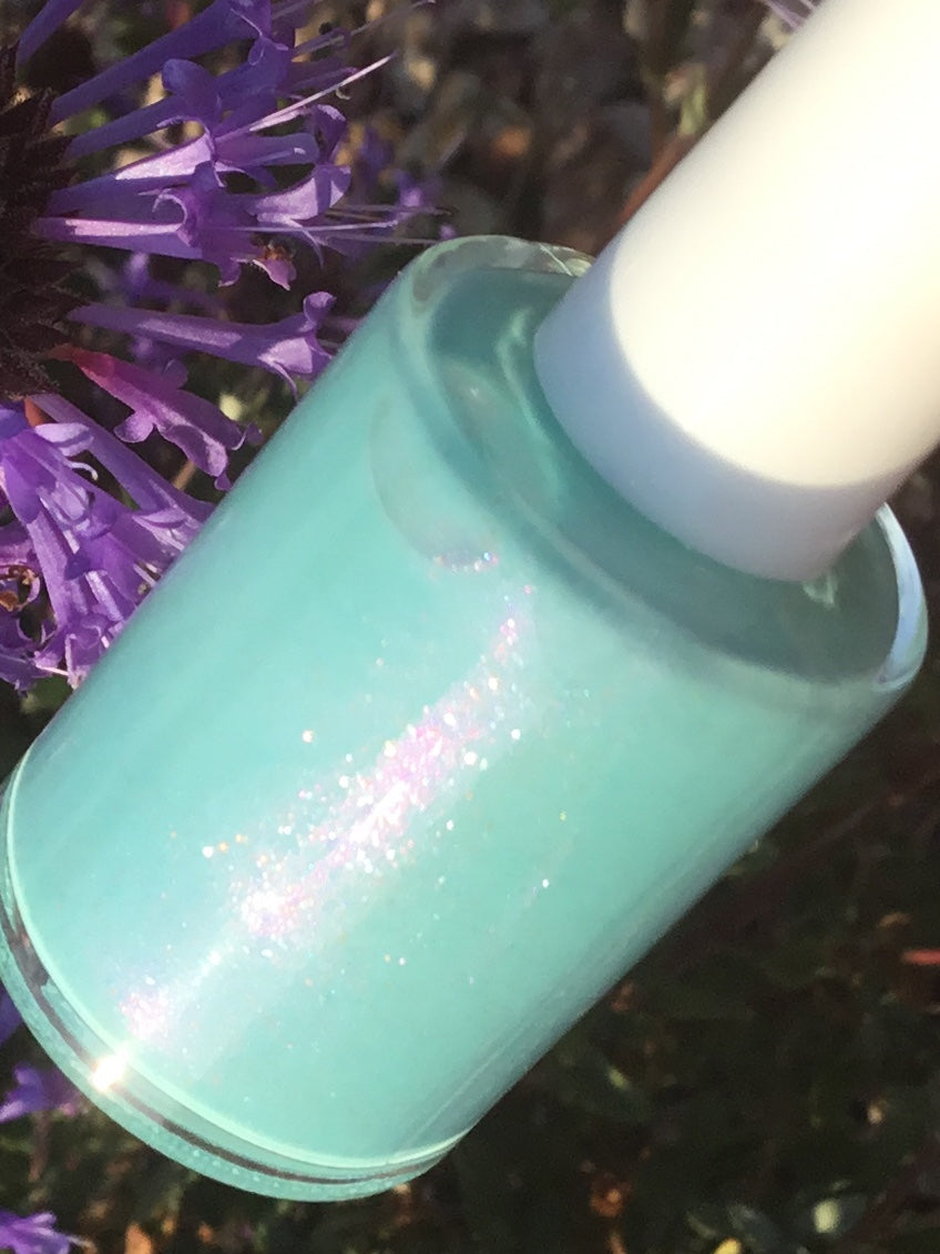 A bottle of Marshmellow Dream.001, a pale green nail polish known for its color-changing properties and pearlescent sheen, is displayed against a backdrop of purple flowers and lush greenery. The polish catches the light beautifully, showcasing its shimmer and iridescence—an exquisite offering from PI Colors Luxury Nail Lacquers.