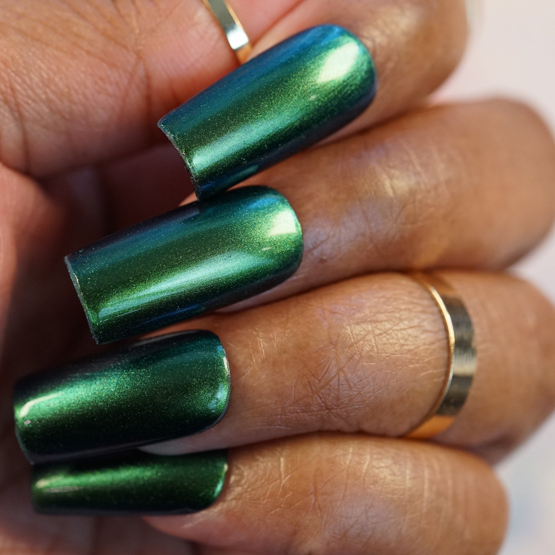 Close-up of a hand with long, square-shaped nails painted in PI Colors' luxurious Green Glow.209 nail polish. The person is wearing gold rings on two fingers, showcasing a modern and stylish manicure.