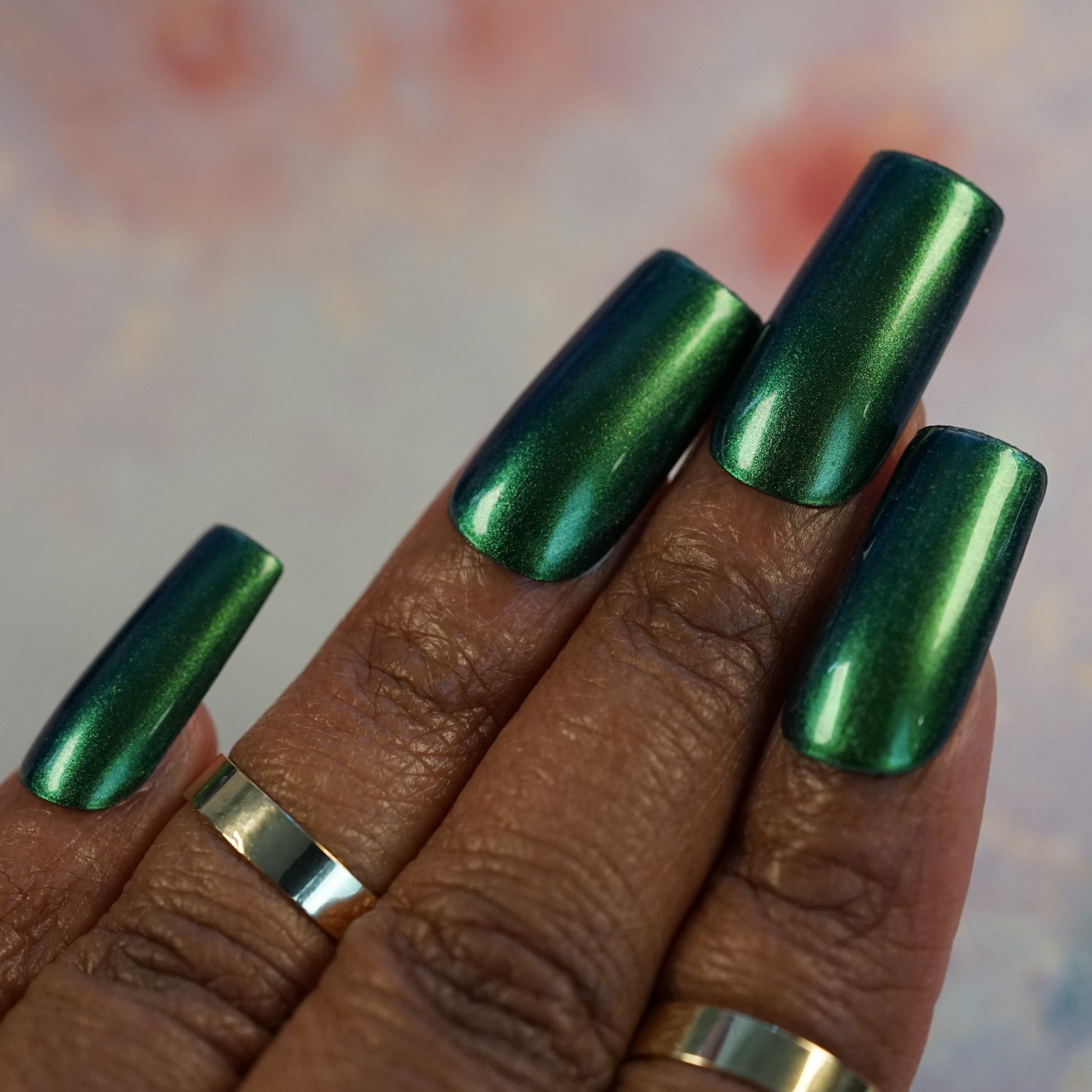A close-up of a hand adorned with long nails painted in Green Glow.209 by PI Colors, showcasing a multichrome green finish with a stunning metallic shine. Luxe gold bands embellish the index, middle, and ring fingers. The blurred background features soft pink and blue hues, further accentuating the brilliance of this luxury nail lacquer.