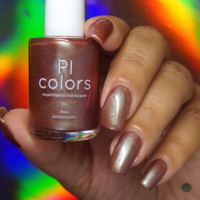 A hand with neatly manicured nails painted in a luxurious rust orange red polish holds a matching Beta Ceti.055 nail polish bottle against a vibrant, rainbow-colored background. The bottle is labeled "PI Colors.