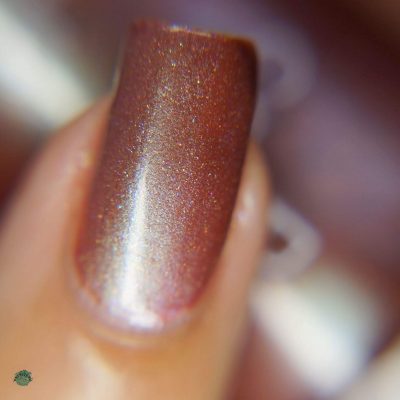 A close-up image showcases a fingernail adorned with PI Colors' Beta Ceti.055 Rust Orange Red Nail Polish, radiating a metallic copper hue interlaced with holographic glitter. The nail's surface appears smooth and glossy, catching and reflecting light beautifully against a blurred backdrop that accentuates its shimmering effect.