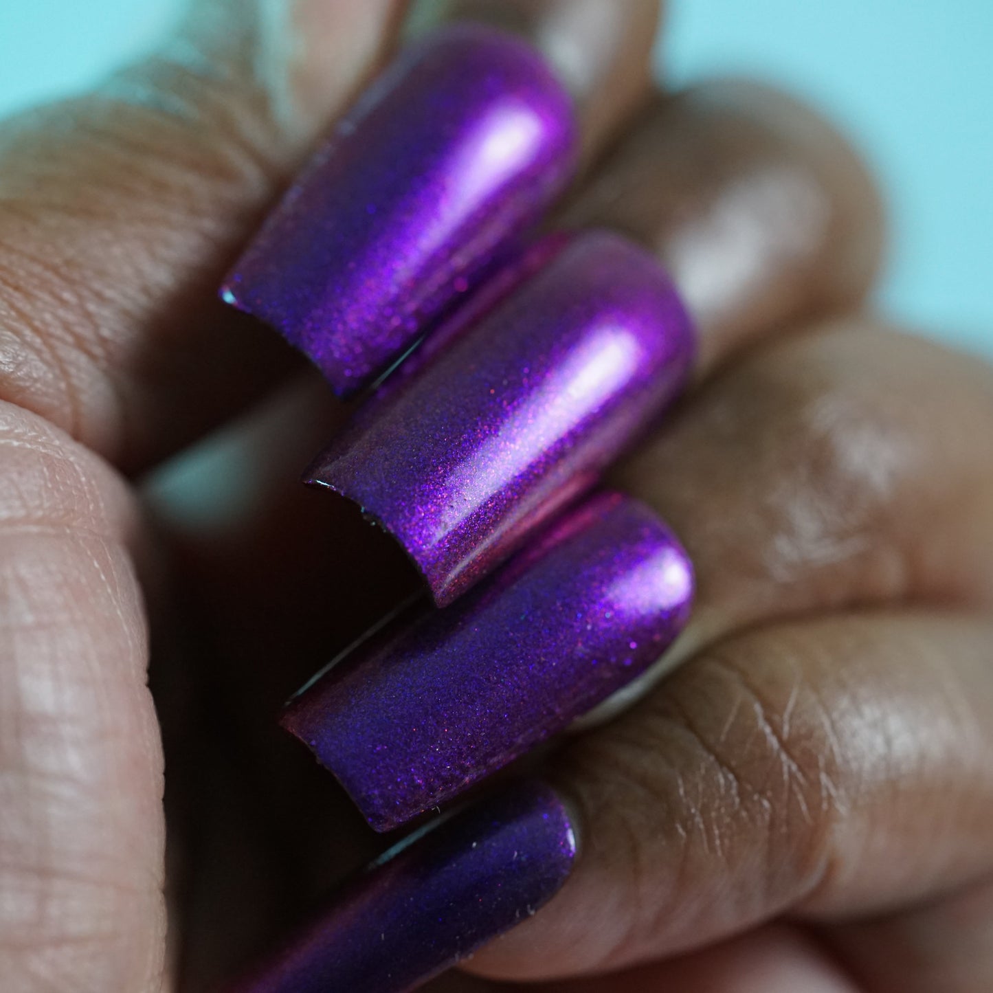 Close-up of a person's hand displaying long, shiny nails painted with Danbury Does It.000 by PI Colors, featuring a purple multichrome. The nails are neatly shaped with a glossy finish against a blurred turquoise background and are 10-free for added peace of mind.