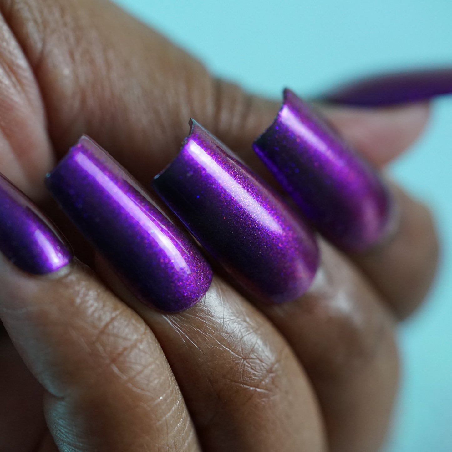 A hand sports long, glossy purple multichrome nails from the PI Colors Danbury Does It.000 collection. The manicured fingers are elegantly curled against a softly blurred backdrop, showcasing the vibrant 10-free polish.