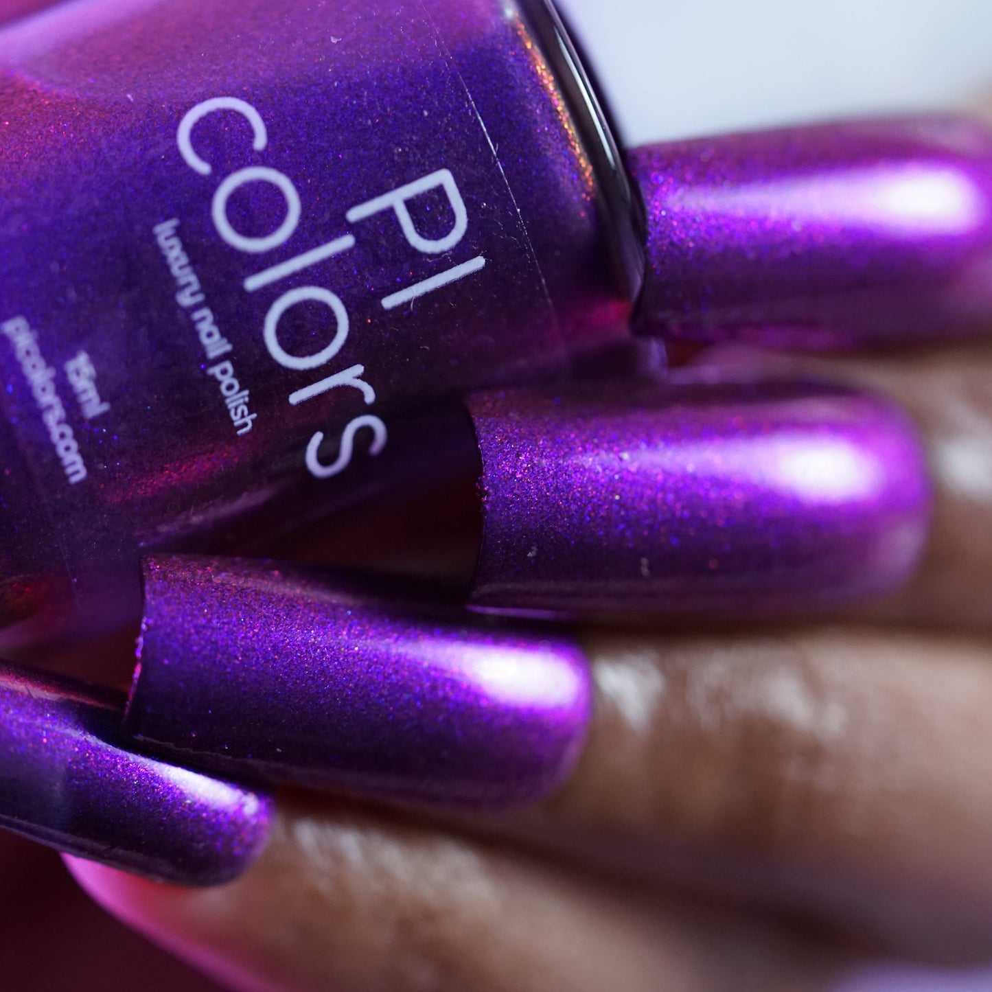 A hand with long, metallic purple multichrome nails holds a matching "Danbury Does It.000" nail polish bottle from PI Colors. This 10-free polish features a glossy finish with shimmering lighter purple hues for an opulent look.