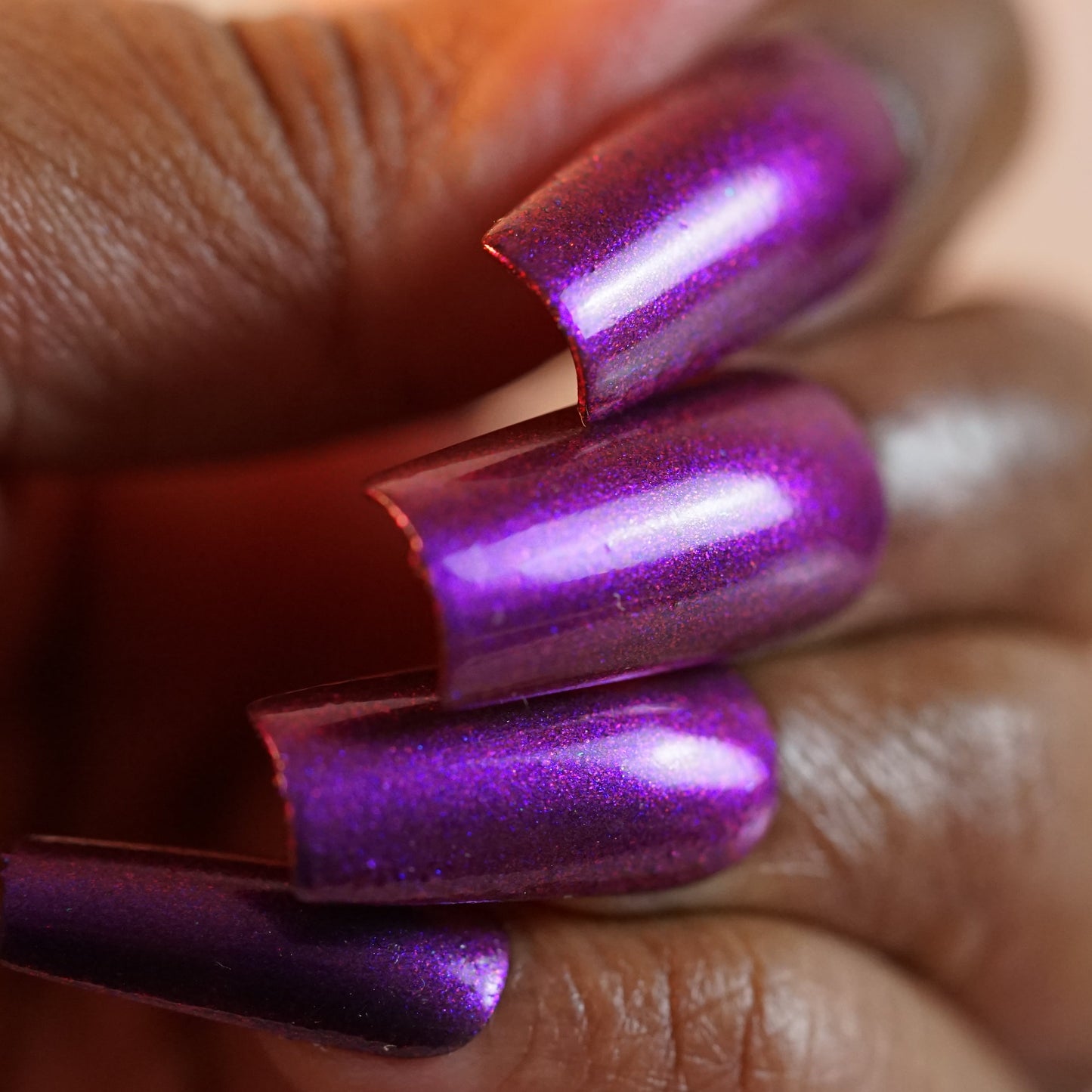 A hand with long, glossy nails in PI Colors’ Danbury Does It.000, a purple multichrome lacquer, reveals slightly squared-off tips and a metallic sheen. The lighting accentuates the vibrant hue and 10-free texture of this luxury polish.